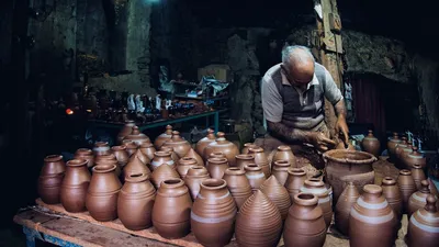 Elderly potter skillfully shapes clay in a dimly lit workshop surrounded by handcrafted pots.