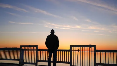 A man stands on a pier, silhouetted against a stunning sunset over calm waters.