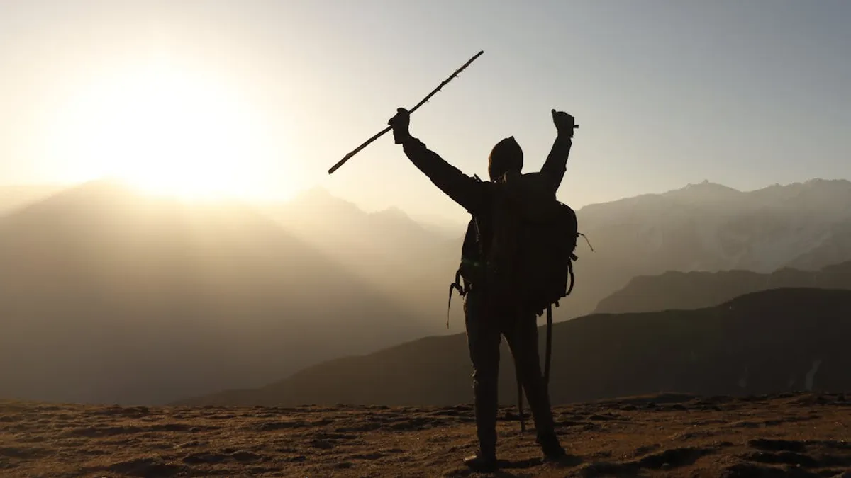 A lone backpacker raises arms triumphantly at sunrise on a mountain peak.