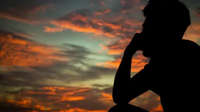 A silhouette of a man sitting and contemplating against a vibrant sunset sky filled with warm hues.