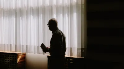 A silhouette of a man standing by a window with sheer curtains in a dimly lit room.