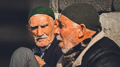 Two senior men in knit caps having a conversation outdoors with warm sunlight.