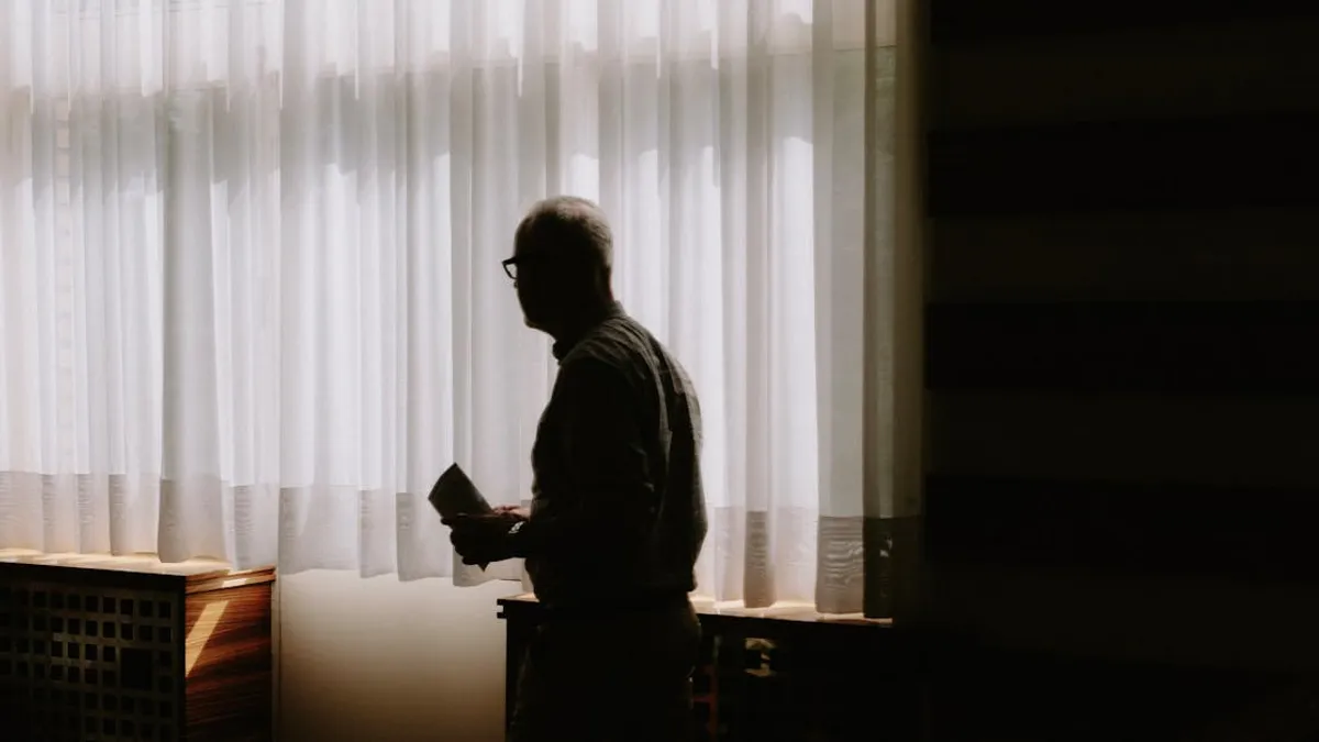 A silhouette of a man standing by a window with sheer curtains in a dimly lit room.