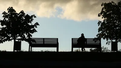 Silhouette of a person sitting on a bench under cloudy skies in a Moscow park at sunset.