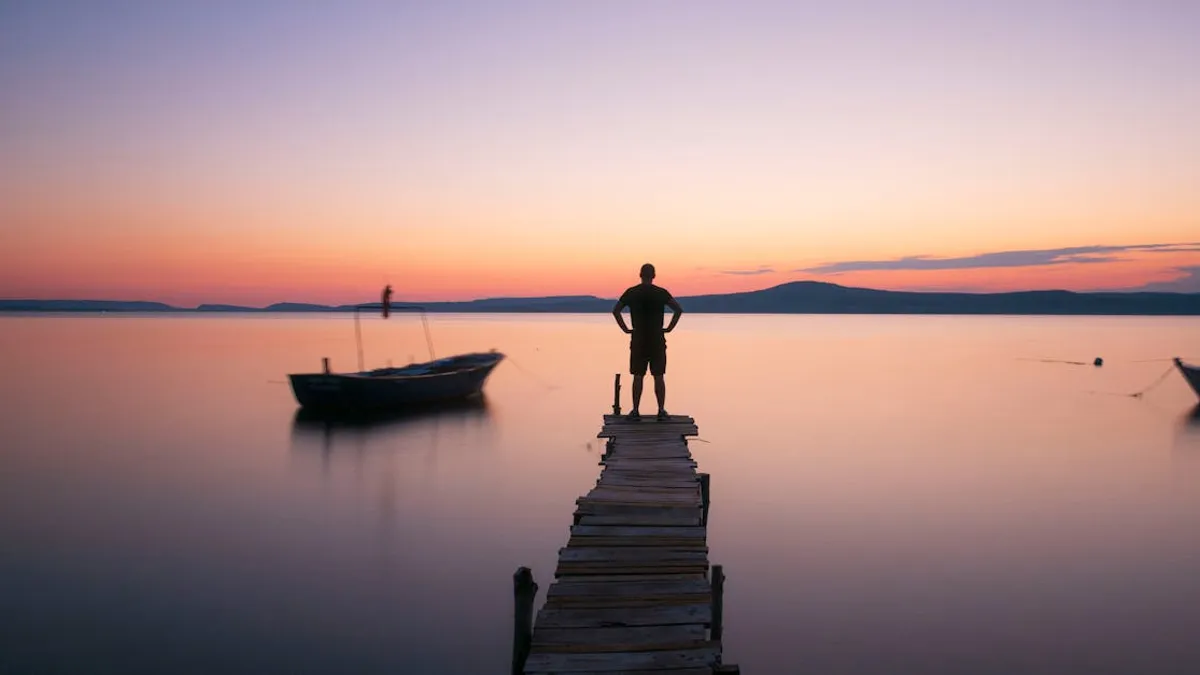 Lonely figure on a wooden pier overlooking calm water during a colorful sunset.