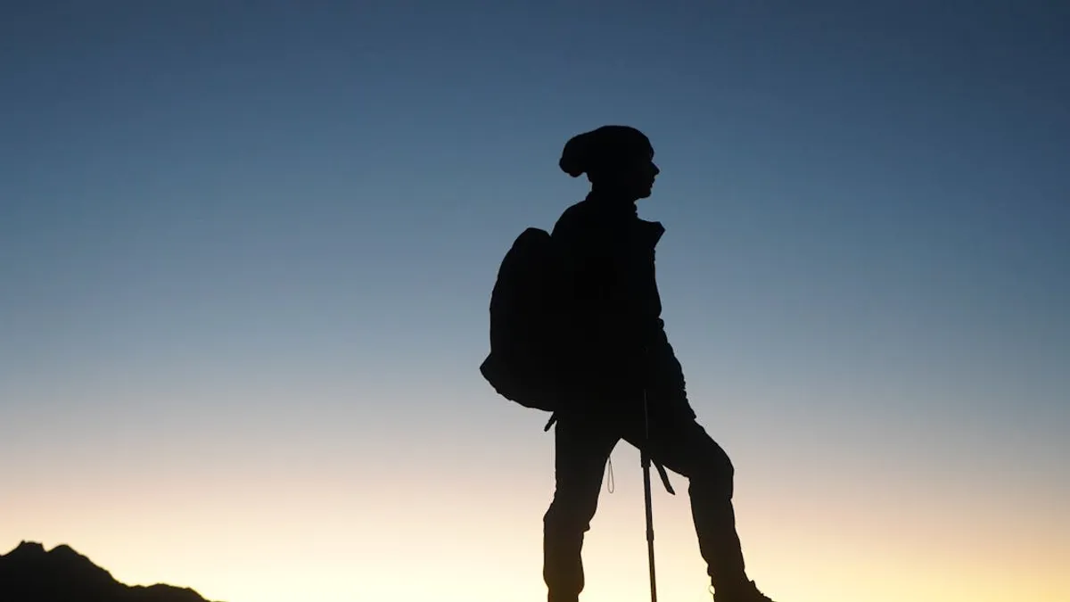 A lone hiker stands silhouetted against a stunning sunrise sky on a mountain peak.