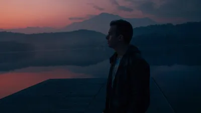 A man in a jacket stands on a dock, gazing at a lake and mountains during twilight.