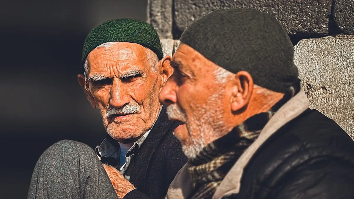 Two senior men in knit caps having a conversation outdoors with warm sunlight.